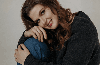 Портрет улыбающейся женщины с каштановыми волосами в сером свитере на нейтральном фоне